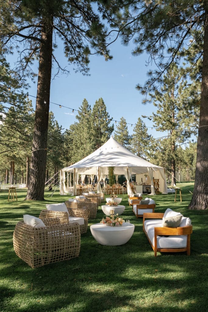 Elegant outdoor wedding reception lounge with a white tent, woven furniture, and contemporary seating beneath towering pine trees.