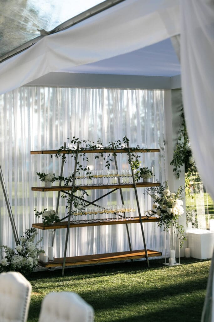 Elegant champagne display featuring gold shelving, white floral arrangements, and sheer drapery beneath a luxury outdoor wedding tent.