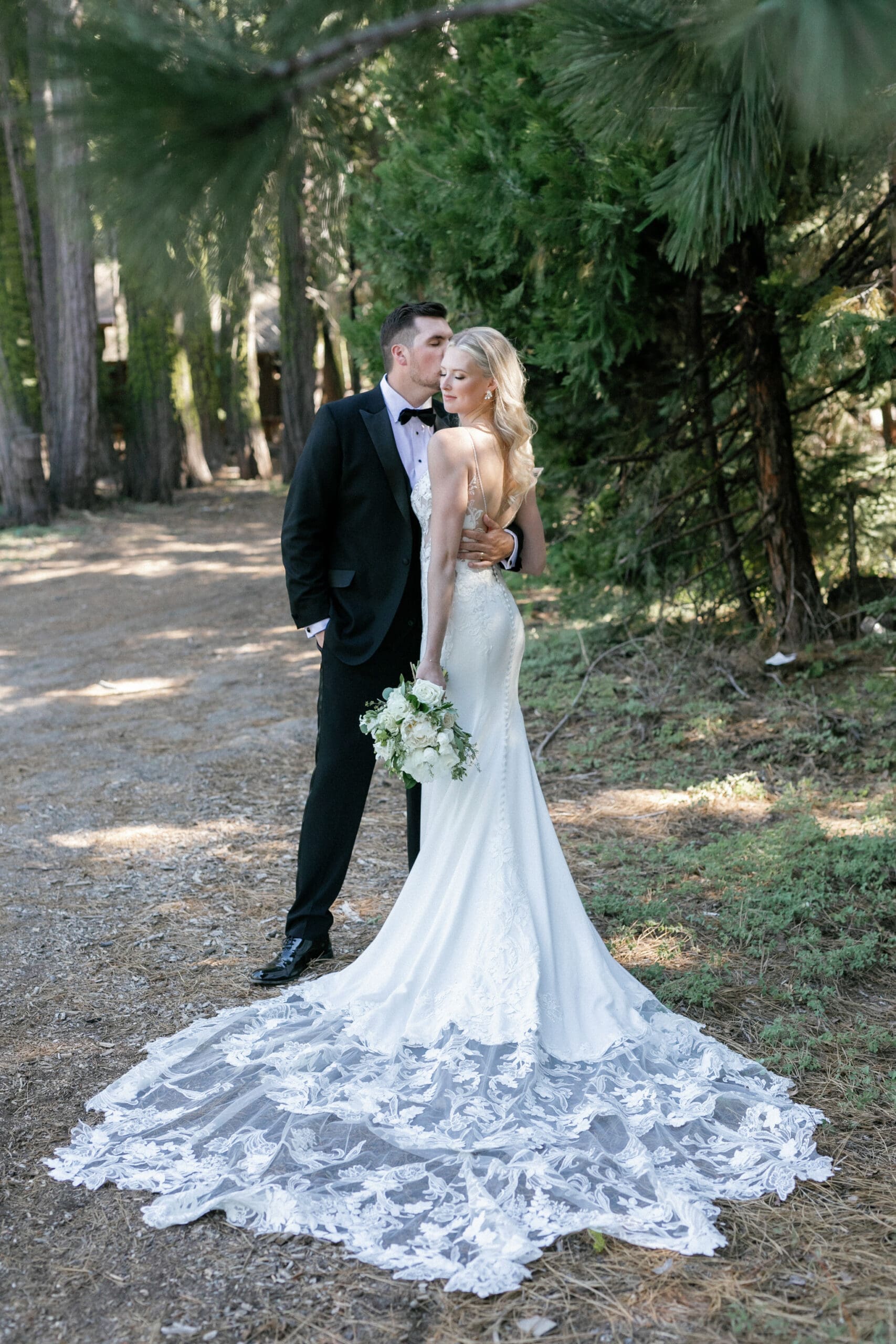 Bride and groom portrait beneath towering pine trees near West Shore Cafe in Lake Tahoe, with the bride’s lace train spread across the forest path at sunset.