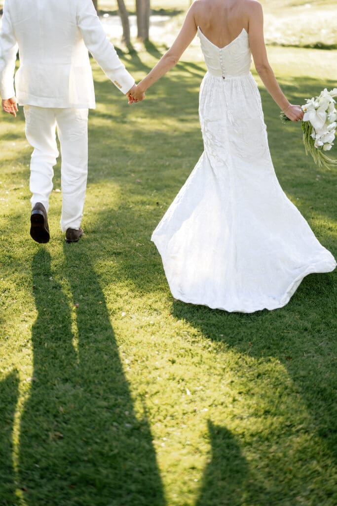 Bride in a fitted white wedding gown holding hands with her groom as they walk across a sunlit lawn during a mountain wedding.