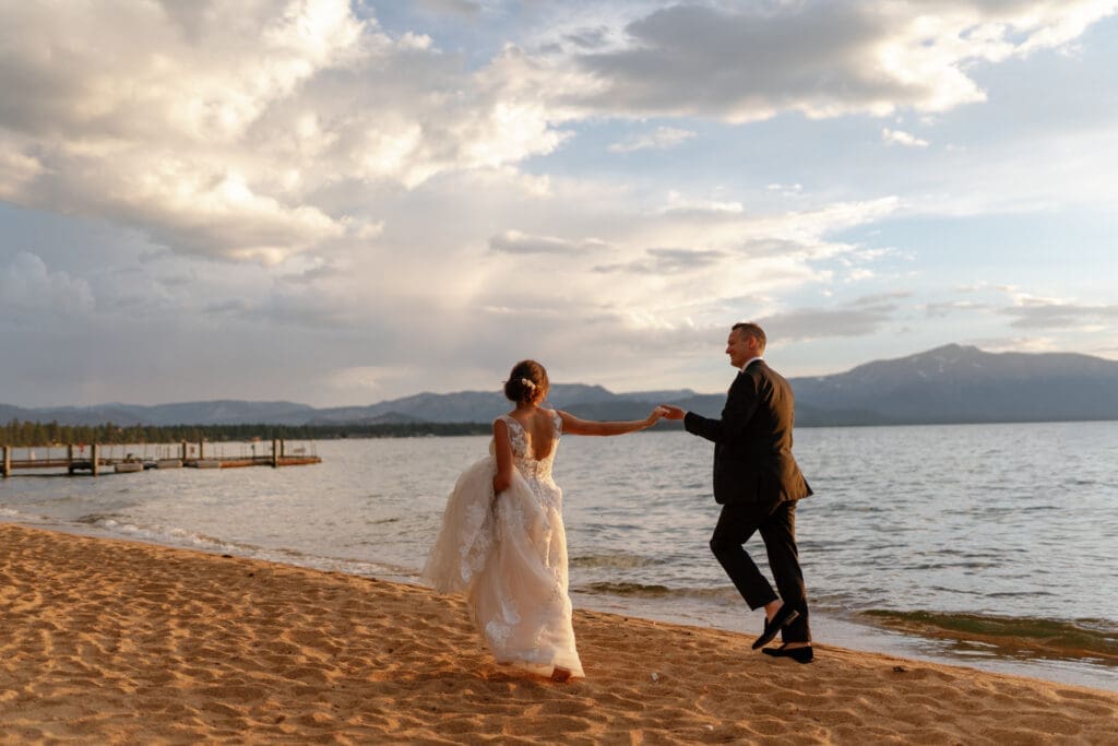 Edgewood Tahoe wedding photography capturing newlyweds holding hands along the sandy shoreline at sunset, with golden light reflecting across Lake Tahoe.