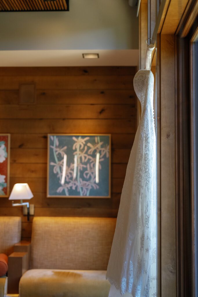 White lace wedding gown hanging beside a window inside a luxury mountain resort suite with warm wood-paneled interiors.