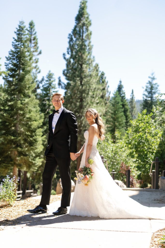Bride and groom holding hands in formal wedding attire surrounded by towering pine trees at a luxury Lake Tahoe wedding venue.