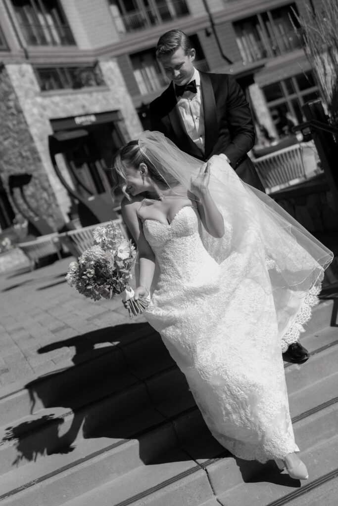 Black-and-white photograph of a groom holding his bride's flowing veil as they walk together outside a luxury mountain resort.