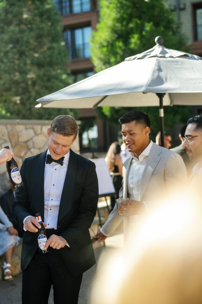 Groom in a black tuxedo opening a drink during an outdoor wedding cocktail hour with guests at a luxury mountain resort.