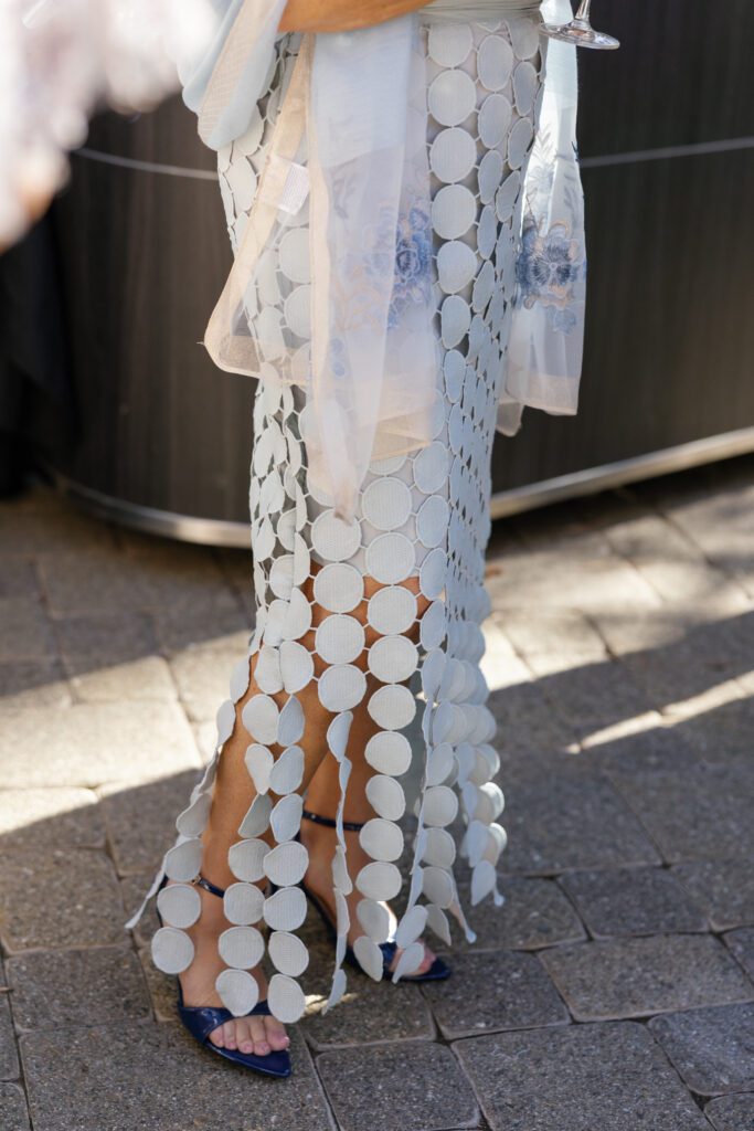 Close-up of a wedding guest wearing an elegant white circular appliqué dress with sheer fabric and navy blue heels at a luxury wedding celebration.