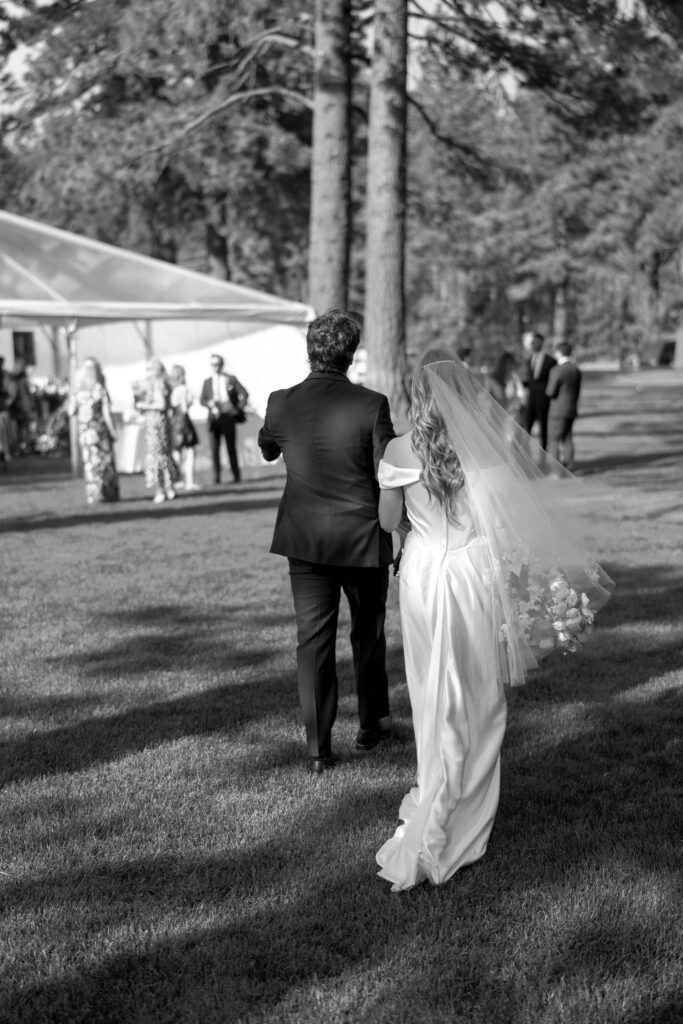 Black-and-white photograph of newlyweds walking across a lawn toward an outdoor wedding reception tent surrounded by pine trees.