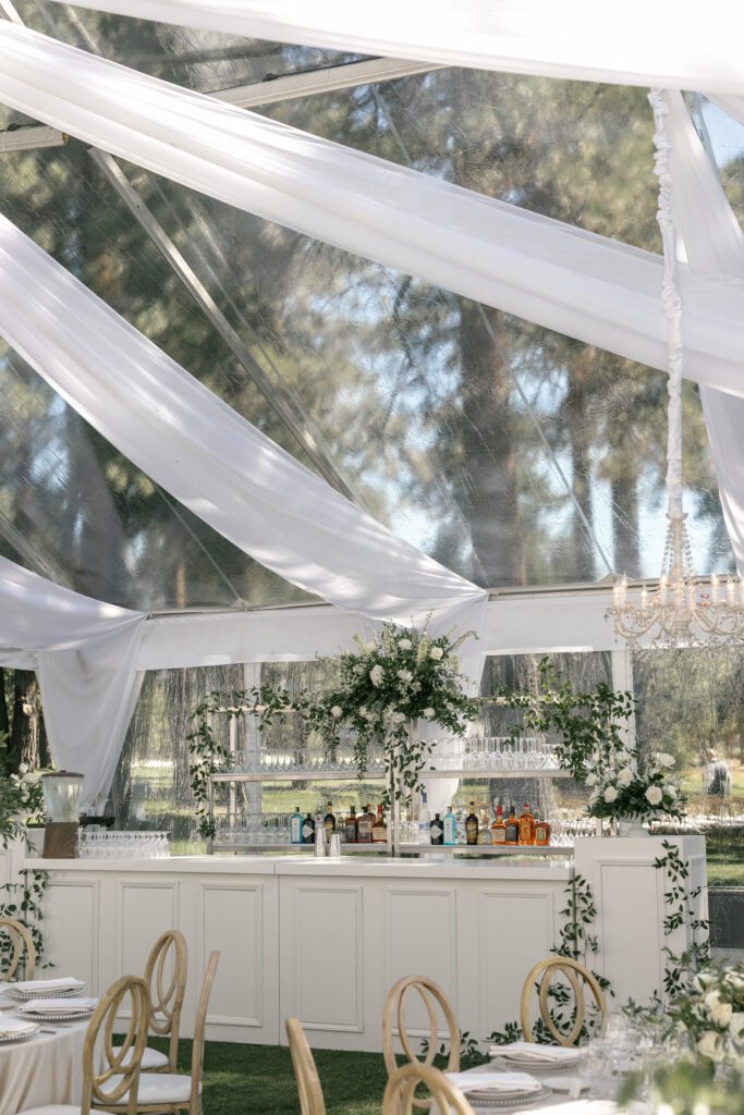 Sophisticated wedding reception bar beneath a transparent tent with flowing white drapery, crystal chandeliers, and lush floral arrangements.