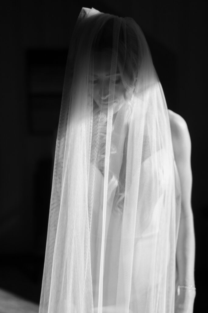 Black-and-white editorial portrait of a bride wearing a sheer wedding veil against a dark background.