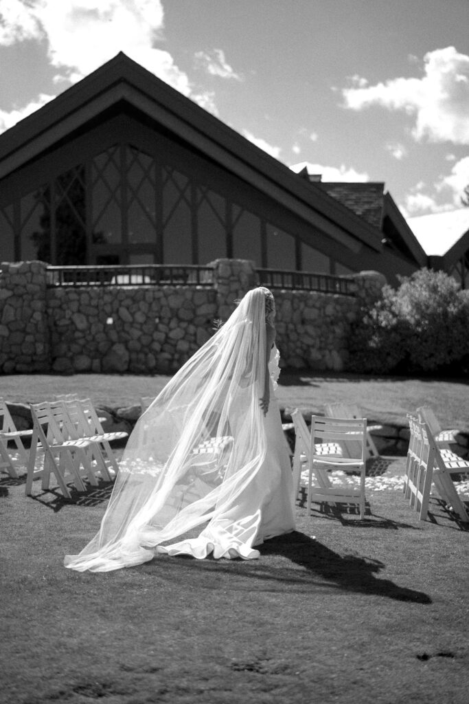 Black-and-white photograph of a bride wearing a flowing veil and wedding gown outside a mountain lodge with stone architecture.