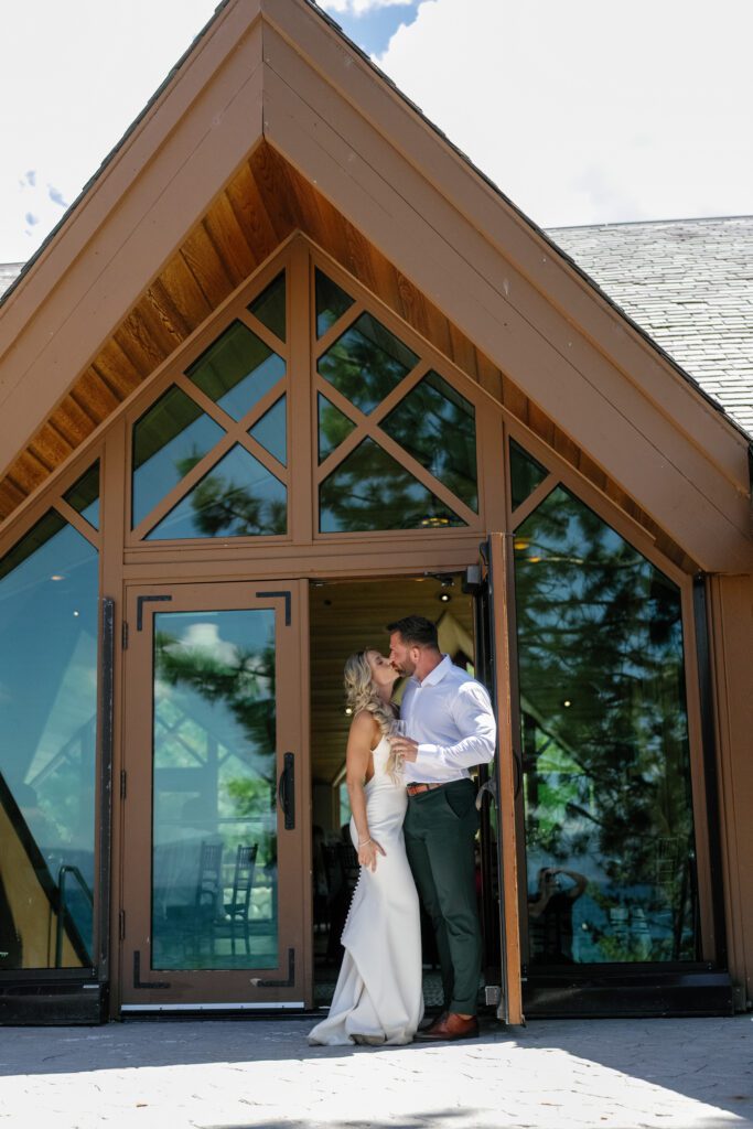 Bride and groom embracing outside a modern A-frame wedding chapel with wood beams and floor-to-ceiling glass windows.