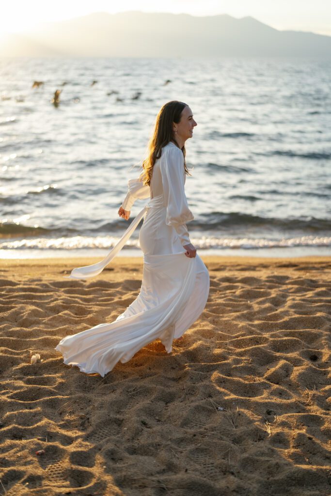 Bride wearing a flowing white wedding gown walking along the sandy shoreline of Lake Tahoe in warm golden-hour light.