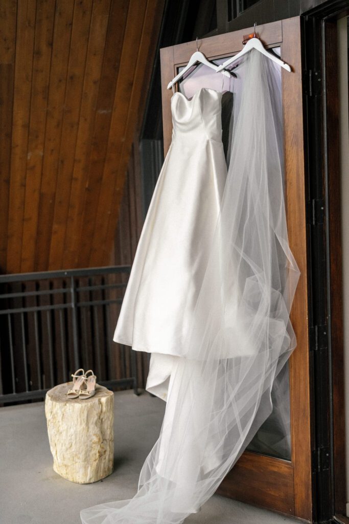 White wedding gown and veil hanging against rustic wood architecture beside bridal accessories displayed on a natural stone pedestal.