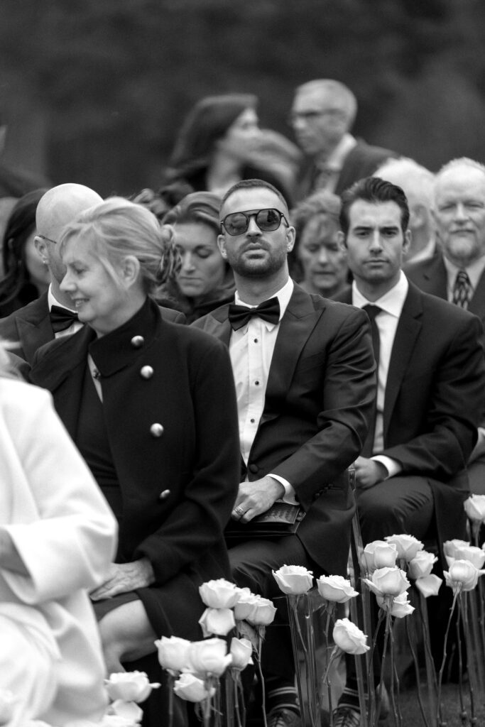 Black-and-white photograph of wedding guests wearing formal tuxedos and elegant attire during a black-tie wedding ceremony.