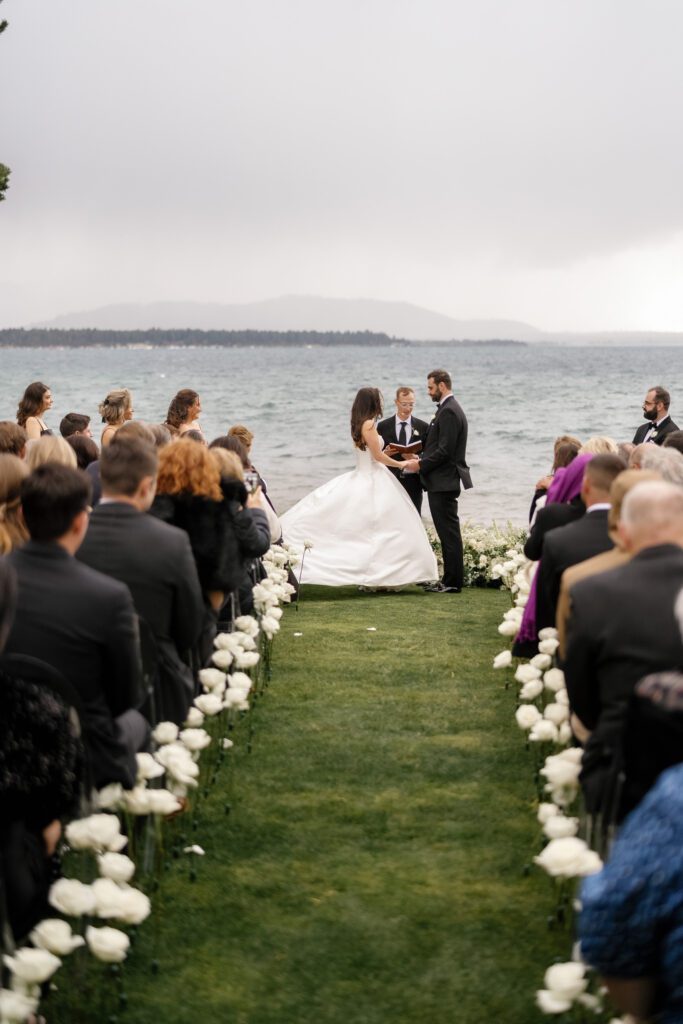 Bride and groom exchanging vows during an Edgewood Tahoe wedding ceremony overlooking Lake Tahoe, with white floral arrangements lining the aisle.