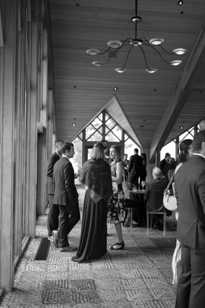 Black-and-white photograph of wedding guests gathering inside an elegant mountain lodge with vaulted ceilings and large windows.