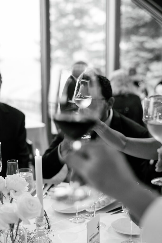 Black-and-white photograph of wedding guests raising champagne glasses during an intimate reception dinner with white floral arrangements.