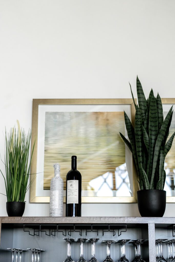 Minimalist wedding bar featuring a framed landscape painting, wine bottle, and contemporary greenery against a white wall.