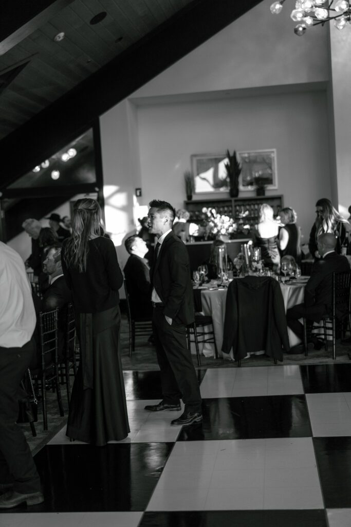 Black-and-white photograph of an elegant wedding reception with guests mingling beneath vaulted ceilings and chandeliers.