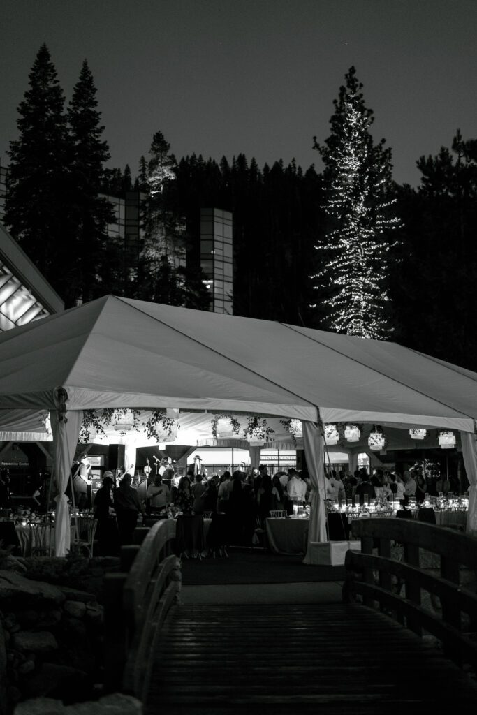 Black-and-white photograph of an outdoor wedding reception tent illuminated at night beneath towering pine trees in Olympic Valley.