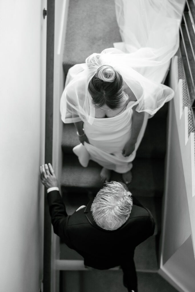 Black-and-white overhead photograph of a bride in a flowing wedding gown meeting an older gentleman on a narrow staircase.