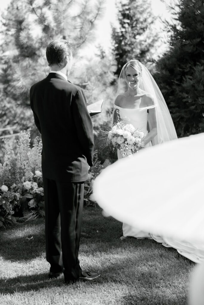 Black-and-white photograph of a bride and groom sharing an intimate moment during an outdoor wedding ceremony surrounded by trees.