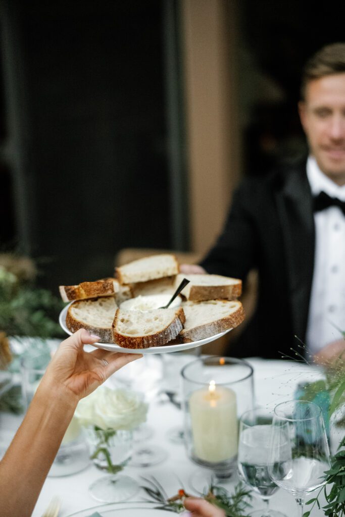 Wedding guest serving rustic bread during an intimate reception dinner featuring candlelight, white floral arrangements, and elegant table settings.