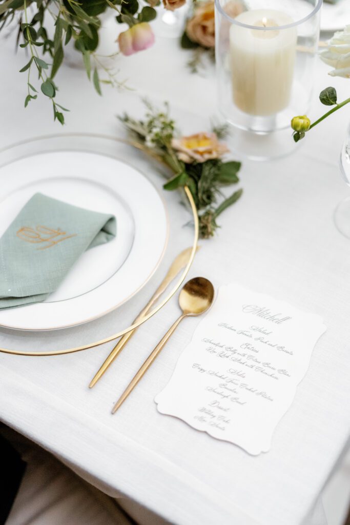 Refined wedding reception tablescape featuring white linens, gold flatware, a sage green napkin, and delicate floral details.