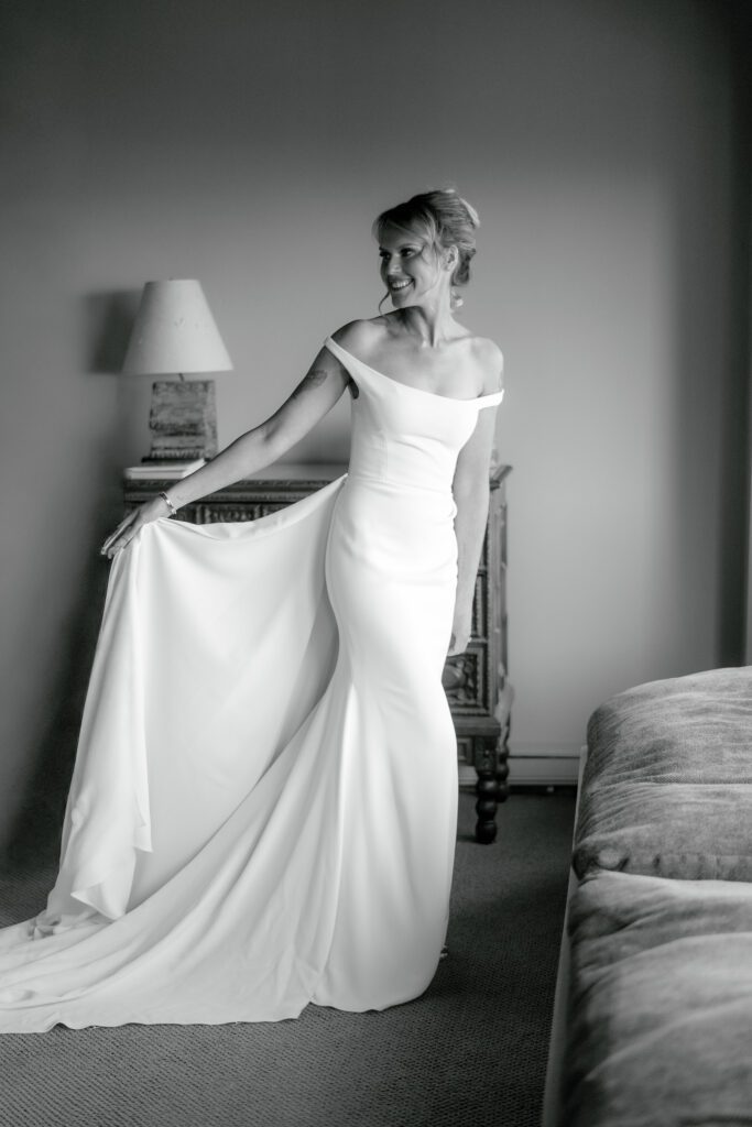 Black-and-white editorial bridal portrait featuring a bride in an elegant off-the-shoulder wedding gown holding her flowing skirt beside a bedroom window.