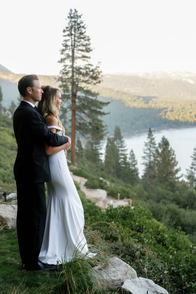 Bride and groom embracing on a mountainside overlooking Lake Tahoe, surrounded by pine trees and the Sierra Nevada landscape.