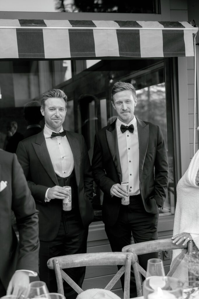 Black-and-white portrait of two wedding guests wearing classic black tuxedos and holding drinks during an elegant private estate reception.
