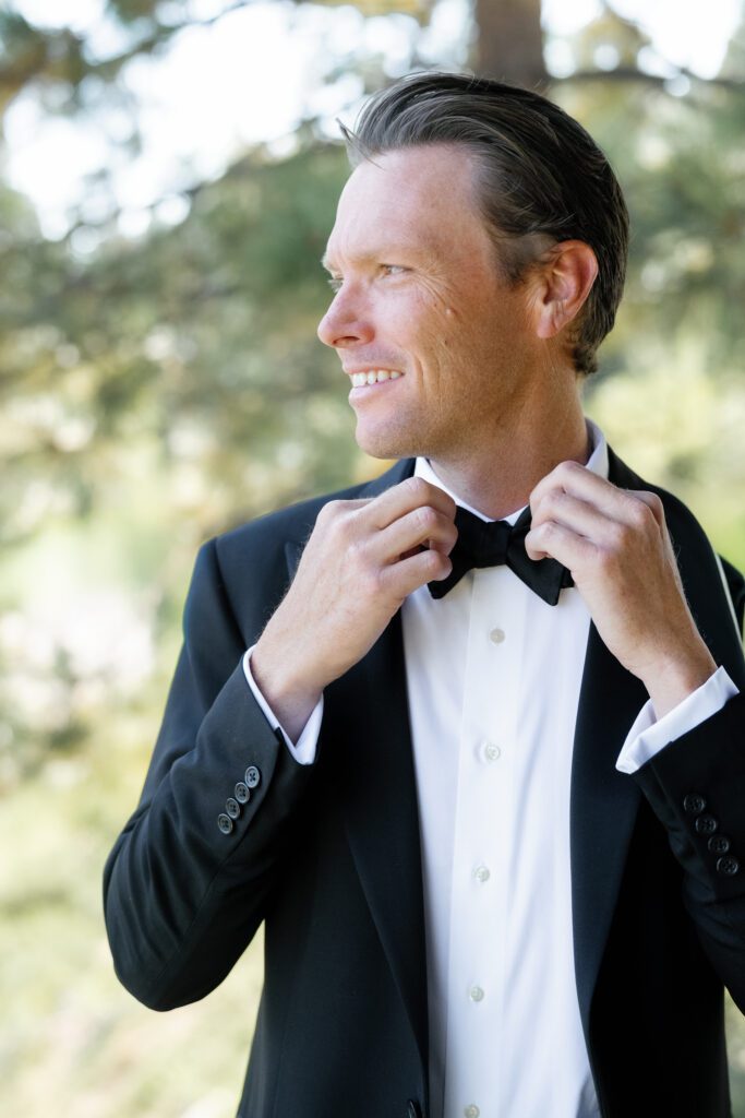 Groom wearing a classic black tuxedo adjusting his bow tie outdoors with soft natural light and greenery in the background.