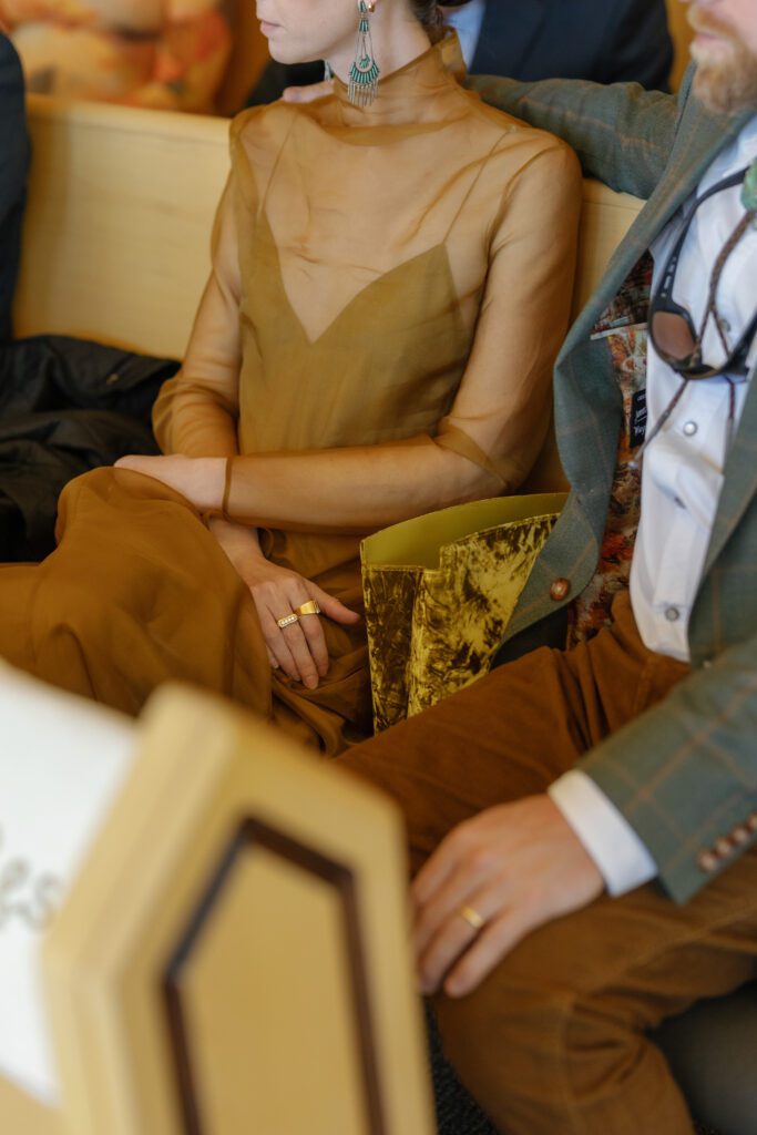 Wedding guest wearing a sophisticated golden-brown dress and statement earrings while seated during an intimate autumn celebration.