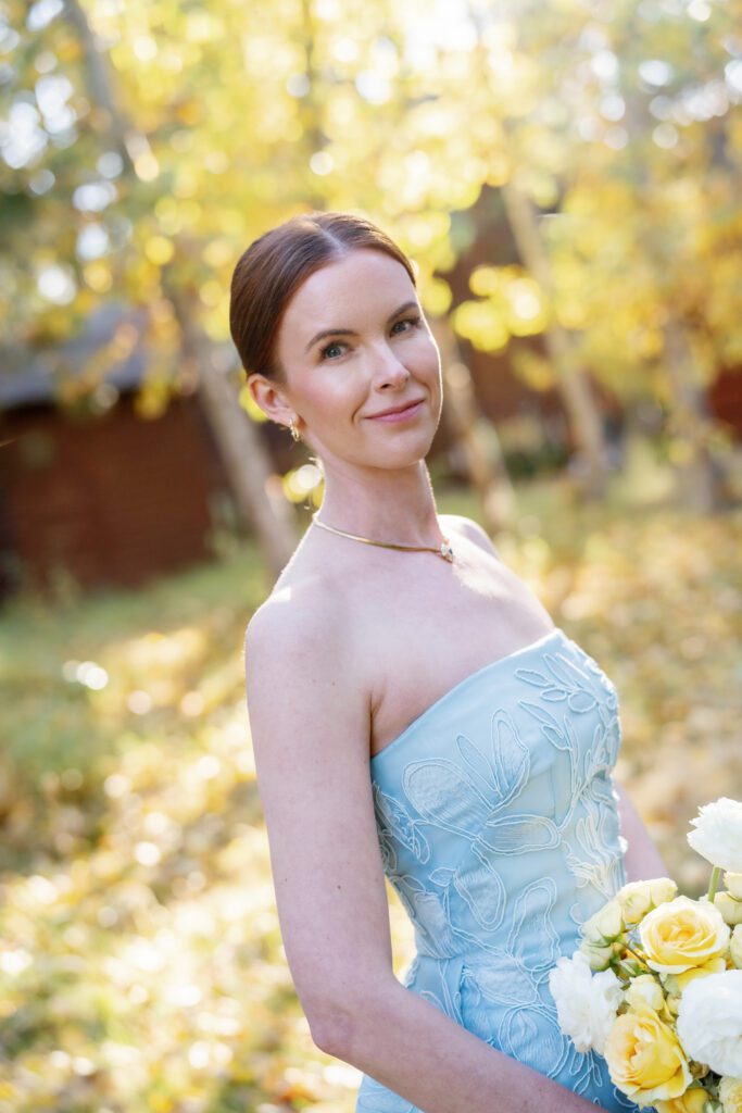 Bride wearing a pale blue strapless gown and holding yellow roses against a backdrop of golden autumn foliage.