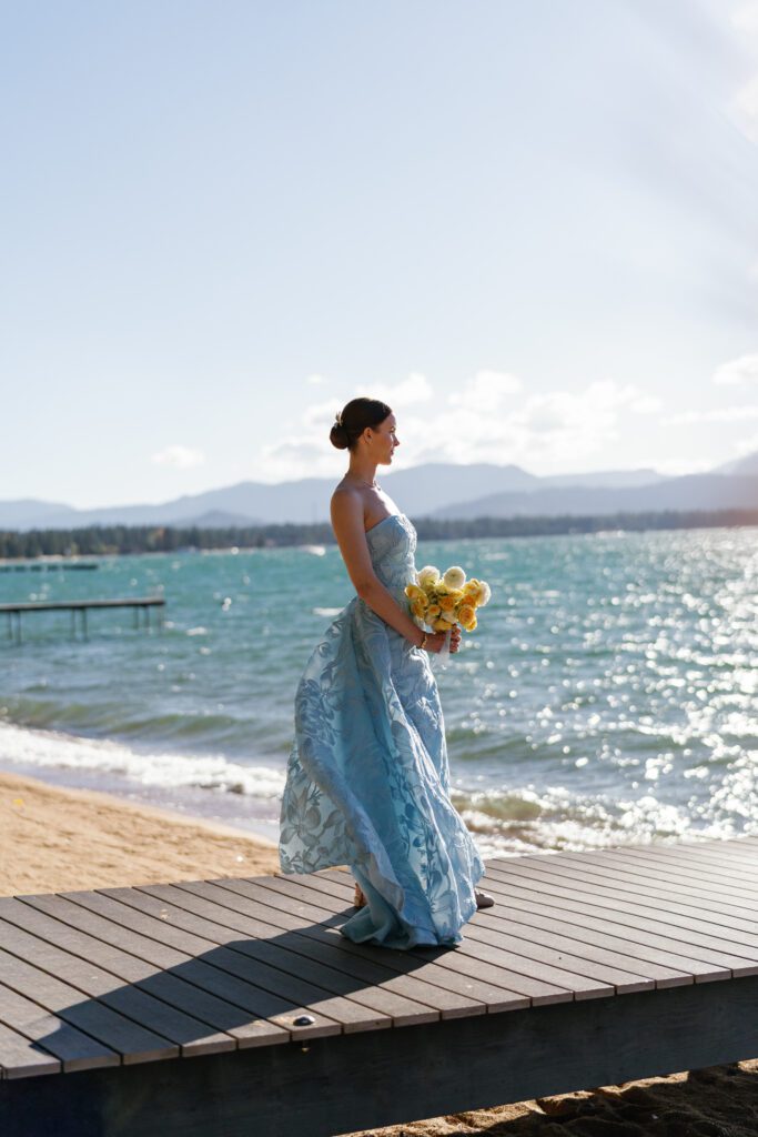 Bride in a blue wedding gown holding a yellow floral bouquet on a wooden dock overlooking Lake Tahoe and the Sierra Nevada mountains.