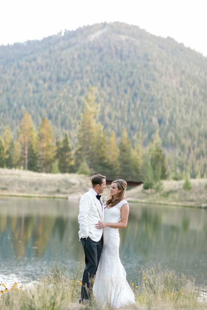 Bride and groom embracing beside a peaceful alpine lake surrounded by forested Sierra Nevada mountains.