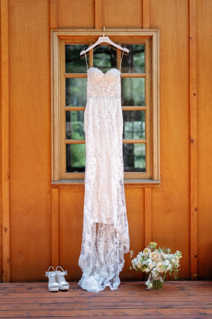 Delicate lace wedding gown hanging against a wooden cabin exterior beside bridal shoes and a colorful floral bouquet.