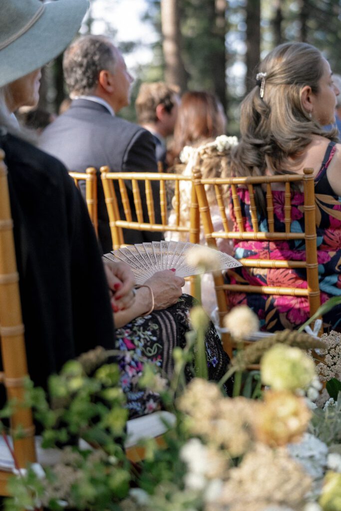 Guests seated on gold chairs beneath towering pine trees during an outdoor Lake Tahoe wedding ceremony with seasonal floral arrangements.